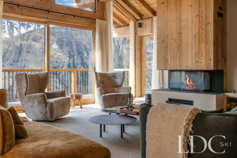 Living room at Chalet Lenafaju with fireplace, armchairs and large windows overlooking the mountains